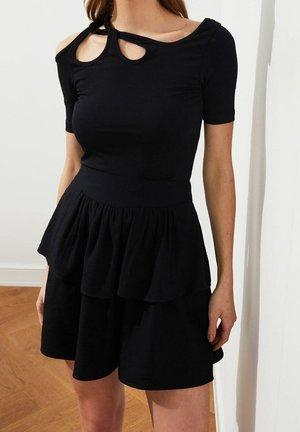 Kvinde iført sort kortærmet kjole med lagdelt flæsenederdel og asymmetrisk halsudskæring med to udskæringer bundet med stofknude.