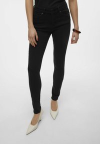 Jeans skinny negros con una textura suave y un diseño de cinco bolsillos. Combinados con tacones blancos de punta, mostrando un estilo minimalista.