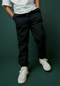 Schwarze Cargo-Hosen mit elastischen Bündchen und Taschen, kombiniert mit weißen Sneakern. Das Outfit hat eine lässige Passform und eine matte Stofftextur.