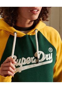 Felpa verde e gialla con cappuccio e cordini, caratterizzata dal logo bianco "SuperDry". Tessuto morbido con design a blocchi di colore e polsini a costine.