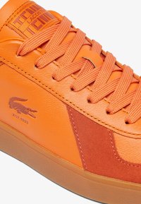 Baskets orange avec une tige en cuir lisse agrémentée de détails en suède, des lacets ronds, et une semelle extérieure en caoutchouc ; logo Lacoste embossé sur le côté.
