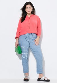Top texturizado coral, con capucha y cordones en la parte delantera; pantalones estampados azul claro con cinturón de lazo; bolso verde; sandalias plateadas con suelas negras.
