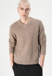 V-neck knitted sweater in light brown, featuring a textured pattern and ribbed cuffs and hem. Model wears it with relaxed-fit black pants.
