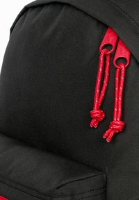 Black fabric backpack featuring bright red drawstring toggles and cords, with a textured surface and a padded bottom area.