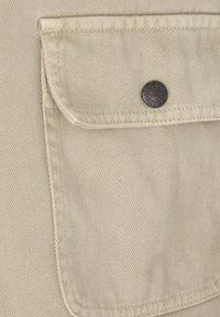 Beige stoflomme med en rund flap og en metal trykknap, som har synlige sting og en subtil diagonal struktur.