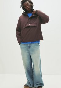 Brun pulloverjacka med ficka framtill, bärs över en blå skjorta, kombinerad med ljusblå baggy jeans och bruna skor. Slät tygkvalitet.
