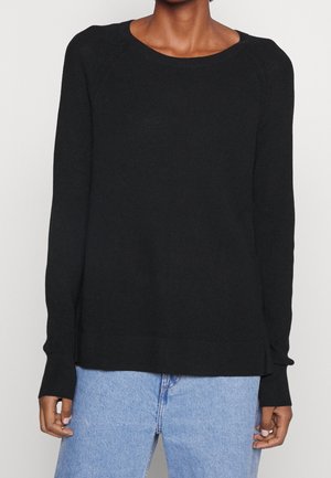 Person wearing a black textured long-sleeve sweater with a round neckline and light blue denim jeans, standing against a plain background.