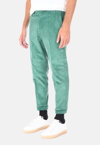 Pantaloni in corduroy verdi con una vestibilità dritta e sottili strisce verticali, dotati di chiusura a bottone e polsini elastici. Abbinati a sneakers bianche.
