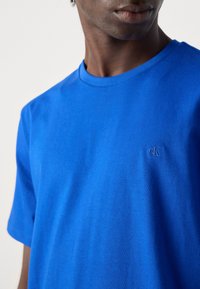 Camiseta de algodón azul con cuello redondo y mangas cortas; pequeño logo bordado en el pecho. Textura suave y corte regular.