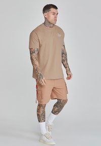 Tan cotton t-shirt with a round neck, paired with tan cargo shorts featuring pockets and red accents. Worn with white socks and sneakers.