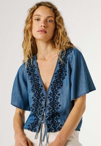 Γυναίκα με κυματιστά ξανθά μαλλιά φοράει μπλε τζιν μπλούζα με μαύρα κεντητά σχέδια, κοντά φαρδιά μανίκια και κλείσιμο μπροστά με δεσίματα.