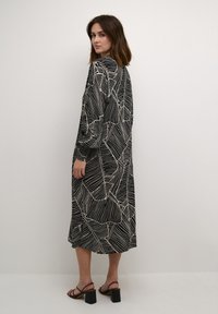 Zwart lange mouwen jurk met beige geometrisch patroon, gemaakt van gladde stof. Met een ontspannen pasvorm en lengte tot halverwege de kuit, gecombineerd met zwarte haksandalen.