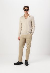 Suéter beige con cremallera hecho de un material suave, combinado con pantalones caqui claros y zapatos negros. El modelo lleva gafas de sol negras.