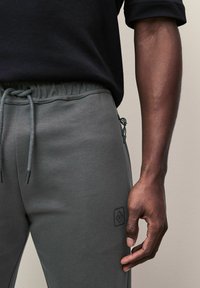 Graue Sweatpants mit einem elastischen Bund und Kordelzug, Seitentasche und einem kleinen schwarzen Emblem am Oberschenkel. Glatte Textur.