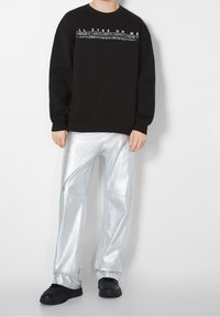 Černá mikina s bílým textem přes hruď, doplněná lesklými stříbrnými kalhotami. Černá obuv dokončuje outfit.