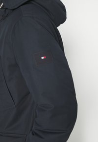 Navy blue jacket with a hood, featuring a patch on the left sleeve with a logo in black, white, and red. Smooth fabric with a matte finish.
