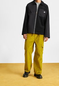 Giacca nera in corduroy con zip e tasca sul petto, abbinata a pantaloni gialli in corduroy dal taglio loose e stivali neri stringati.