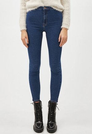 Jean slim bleu porté avec des bottines noires à lacets et un pull crème duveteux, montrés de la taille aux pieds sur un fond uni.