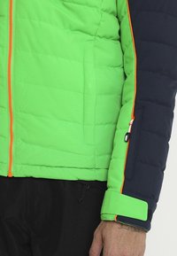Segment d'une veste matelassée verte et bleu marine avec une fermeture éclair orange et des bordures assorties, équipée de poches latérales et d'une fermeture par velcro aux poignets.
