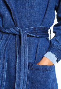 Jeansowy szlafrok w ciemnoniebieskim kolorze, z paskiem do wiązania, bocznymi kieszeniami i prążkowanymi mankietami. Materiał ma teksturowane wykończenie z delikatnym połyskiem.