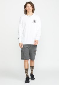 Langärmliges weißes Baumwollhemd mit grafischem Logo, kombiniert mit grauen Cargo-Shorts, gemusterten braunen Socken und schwarzen Turnschuhen.
