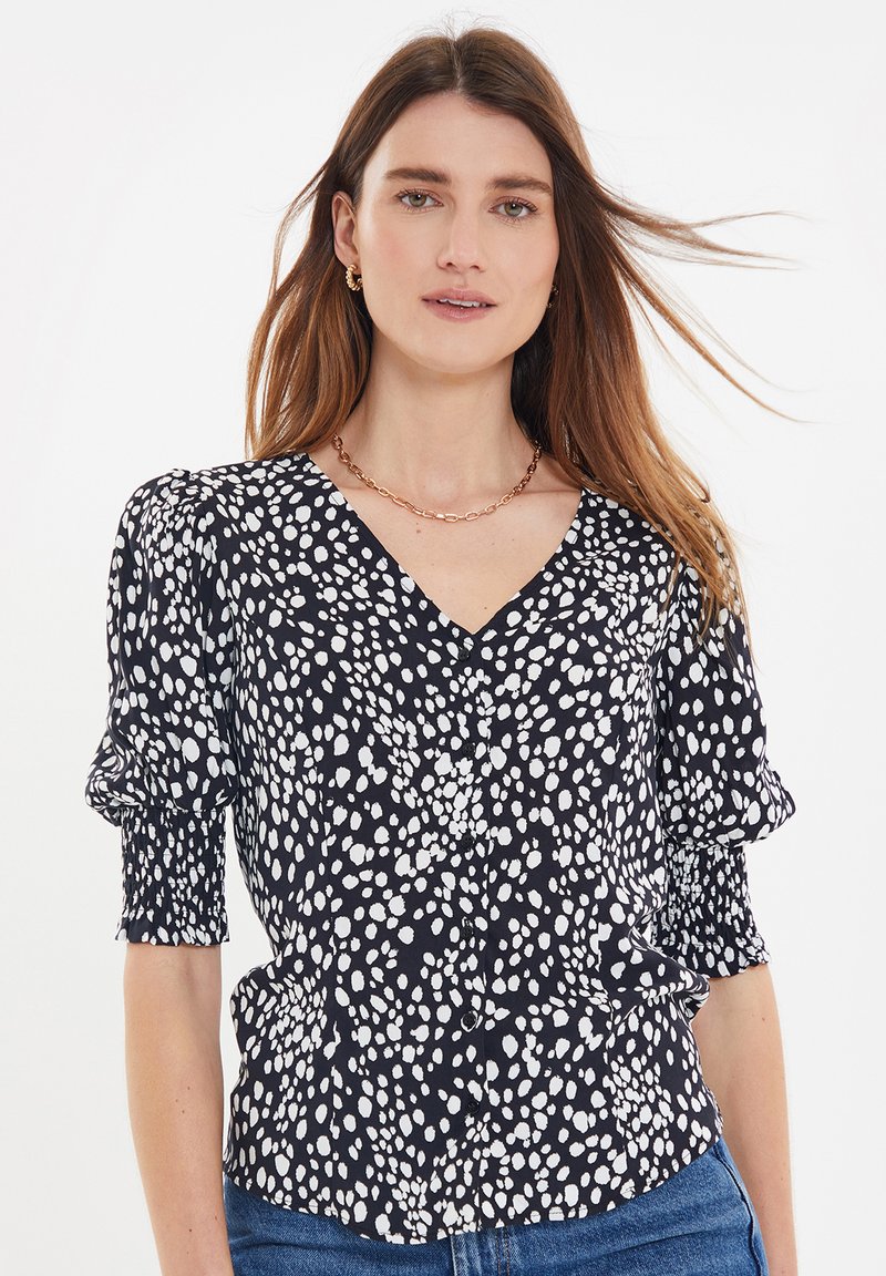 Schwarze Bluse mit weißen Polka-Punkten, ausgestattet mit einem V-Ausschnitt, Puffärmeln und Knopfdetails. Glattes Gewebe mit leichtem Glanz.