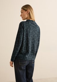 Sudadera verde oscuro con un sutil patrón de leopardo, con mangas largas y cuello redondo, combinada con jeans de mezclilla oscura.