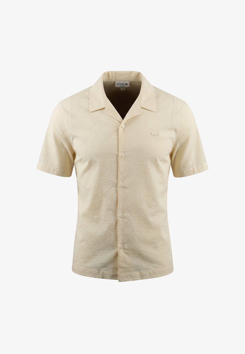 Chemise beige à manches courtes avec boutons, col pointu et tissu subtilement texturé, style unisexe, présentée sur fond blanc.