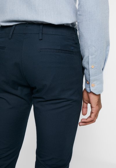 Calças azul-marinho com um design ajustado, feitas de um tecido com um leve brilho. Apresentam um bolso traseiro e um discreto detalhe do logótipo perto da cintura.