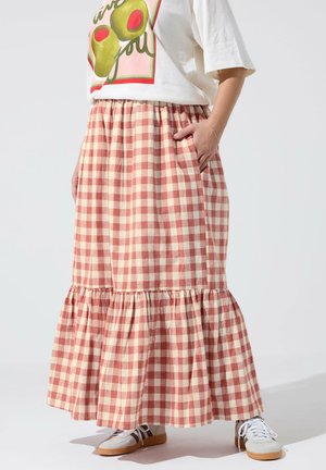 Person iført et langt nederdel i lyserødt og hvidt gingham-mønster, hvid T-shirt med print og hvide sneakers, stående med den ene hånd i lommen.