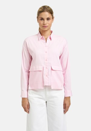 Woman wearing a long-sleeve light pink button-up shirt with two front flap pockets and white pants, standing against a plain background.
