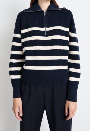 Marineblå sweater med hvide vandrette striber og en halv lynlås foran, båret sammen med matchende marineblå bukser på en stående person.