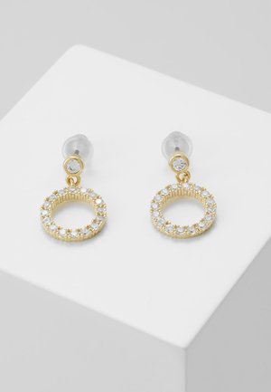 Złote kolczyki koła z małymi przezroczystymi kryształkami wokół koła oraz pojedynczym kryształem przy sztyfcie, na białej skośnej powierzchni.