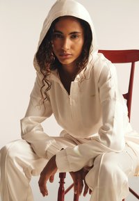 Felpa bianca con zip e vestibilità relaxed, dotata di cappuccio, abbinata a pantaloni coordinati. Tessuto morbido, con polsini elastici e dettagli logo discreti.