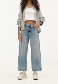 Cardigan grigio chiaro su un top corto bianco, abbinato a jeans a gamba larga blu e sneakers bianche. Nota texture denim sbiadita e vestibilità comoda.