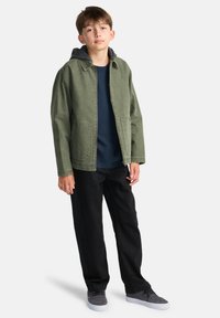 A young person stands confidently, wearing a green jacket over a dark shirt and black pants, with gray shoes, against a white background.