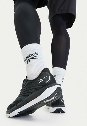 Persona che indossa scarpe da corsa nere Reebok e calzini bianchi con il logo Reebok, che fa un passo avanti su uno sfondo bianco.