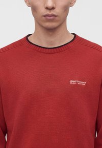 Άνδρας που φοράει κόκκινο πλεκτό πουλόβερ Armani Exchange με ριμπ λαιμόκοψη και λογότυπο στο αριστερό στήθος, φωτογραφημένος από τους ώμους μέχρι το πηγούνι.