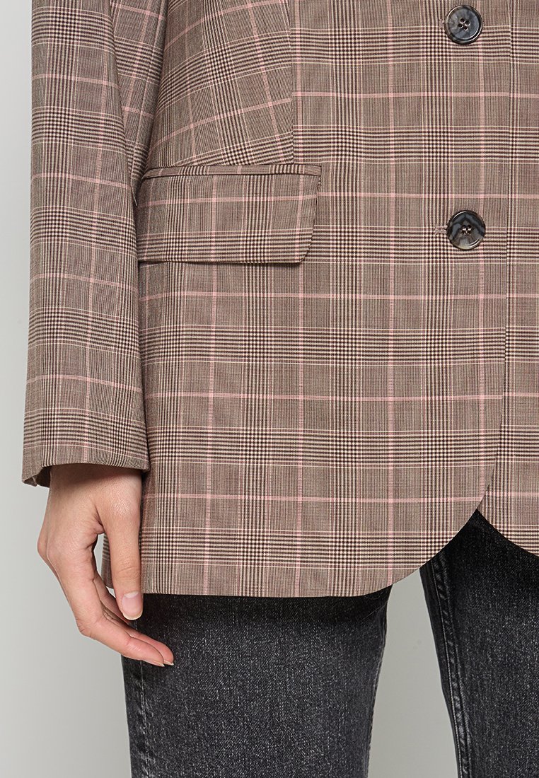 Bruine geruite blazer met roze accenten, voorzien van een geknipte kraag, twee knopen en één zak, gecombineerd met donkere spijkerbroek.