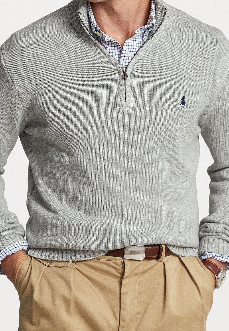 Homme portant un pull gris clair à fermeture éclair partielle sur une chemise à carreaux bleue, un pantalon beige avec une ceinture en cuir marron, mains dans les poches.