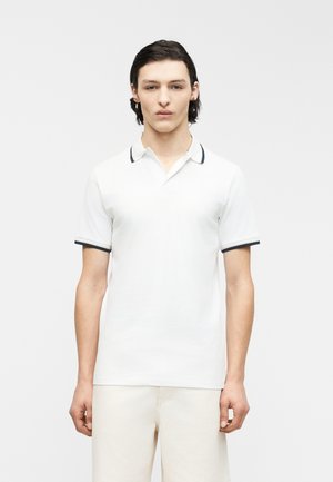 Jeune homme aux cheveux châtains mi-longs portant un polo blanc avec des bordures sombres sur le col et les manches, debout devant un fond uni.