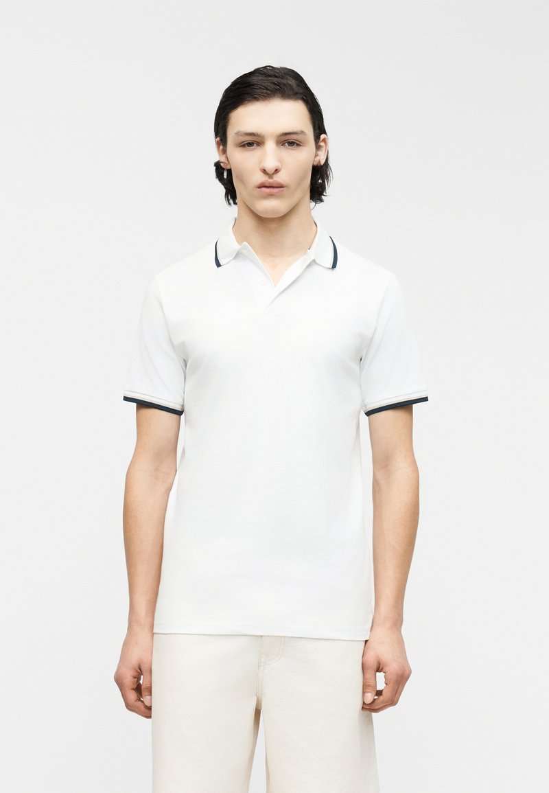 Jeune homme aux cheveux châtains mi-longs portant un polo blanc avec des bordures sombres sur le col et les manches, debout devant un fond uni.