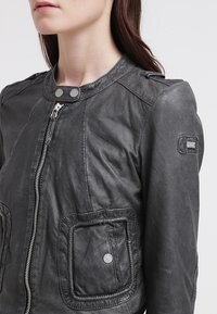 Donna che indossa una giacca di pelle grigio scuro con zip frontale, tasche con bottoni a pressione e una toppa con logo sulla manica superiore, su uno sfondo bianco.