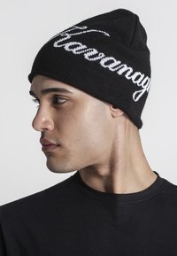 Zwarte gebreide beanie met witte geborduurde letters, slim fit ontwerp en een iets taps toelopende bovenkant. Draagt door een persoon die zijwaarts kijkt.