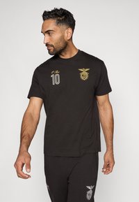 Camiseta de algodón negra con cuello redondo, que presenta un bordado dorado de un emblema y el número "10" en el lado izquierdo del pecho. Diseño simple, con mangas cortas.