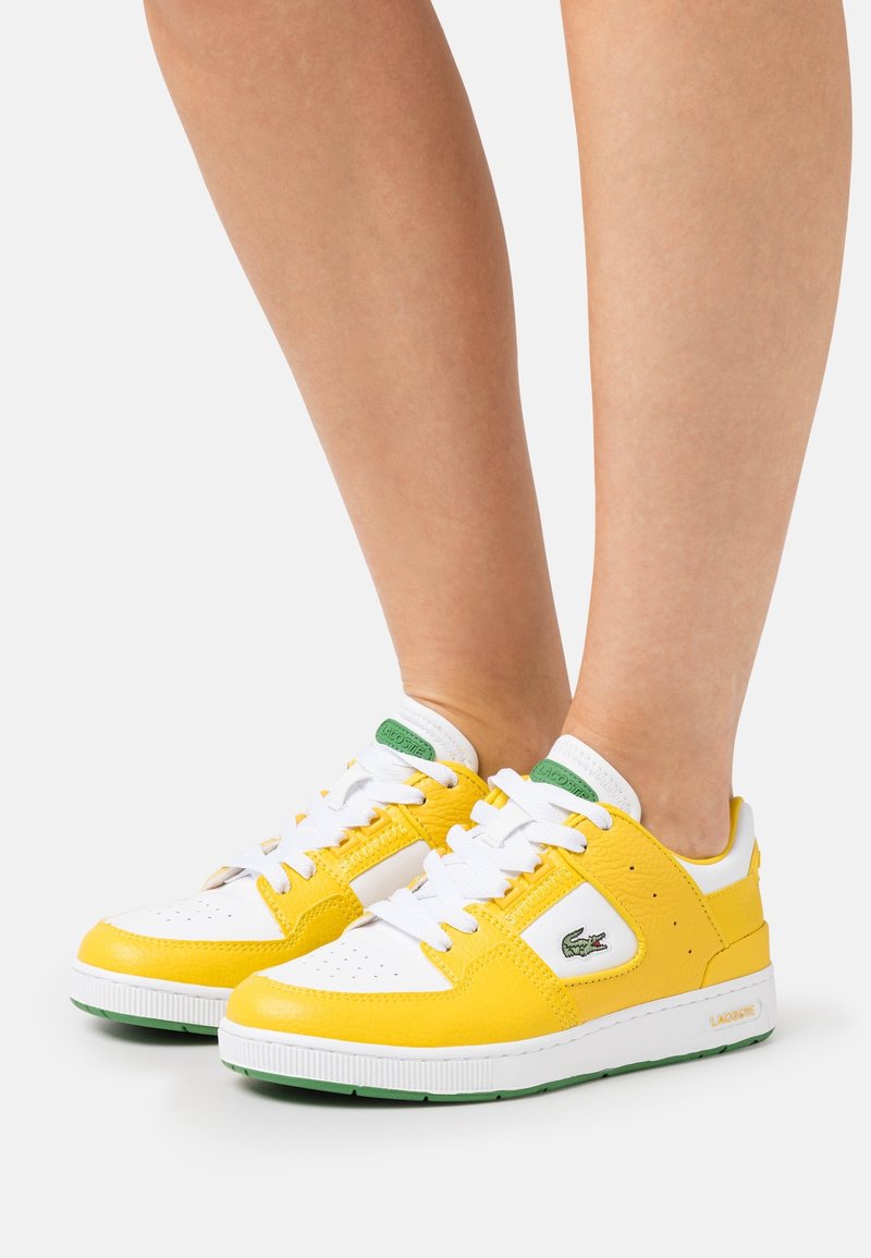 Pieds portant des baskets Lacoste blanches et jaunes avec logo vert, lacets blancs et semelles vertes sur un fond clair.
