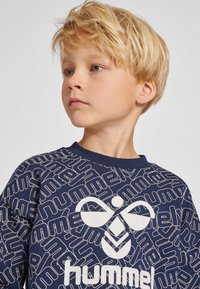Sweatshirt marine avec logo "hummel" blanc et motif répétitif, col rond côtelé, texture douce et manches longues.