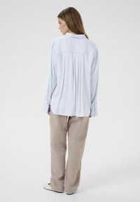 Cămașă albastră deschis cu mânecă lungă, guler și plisuri la spate, combinată cu pantaloni bej largi și pantofi de culoare deschisă.