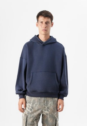 Young man stands against a plain background, wearing a navy hoodie and camouflage cargo pants, hands relaxed at his sides.