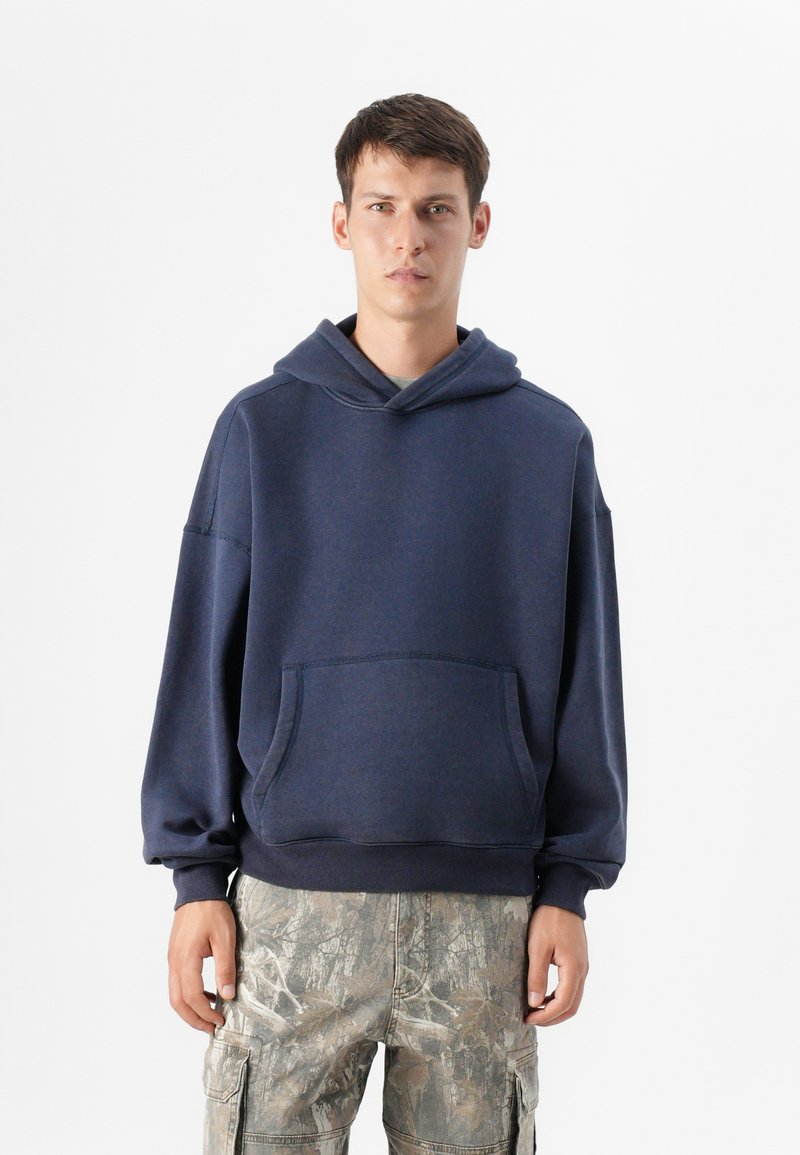 Un tânăr stă în fața unui fundal simplu, purtând un hanorac bleumarin și pantaloni cargo cu model de camuflaj, cu mâinile relaxate la margini.